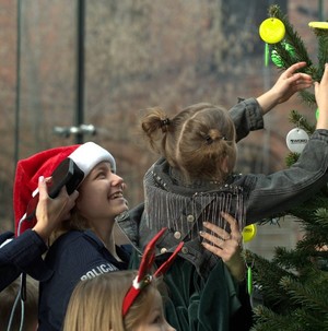 Policjantka w czapce Świętego Mikołaja trzyma a rękach dziewczynkę, która ubiera odblaskową choinkę.