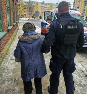 Policjant prowadzi zagubioną seniorkę w stronę karetki.
