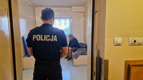 Policjant stoi w drzwiach, w tle widać mężczyznę doprowadzonego do wytrzeźwienia w policyjnej celi.