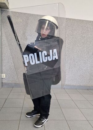 Chłopiec ma założone elementy wyposażenia policjanta biorącego udział w zabezpieczeniach imprez masowych.