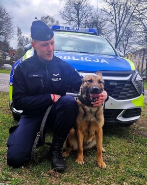 Umundurowany przewodnik z psem na tle oznakowanego radiowozu.