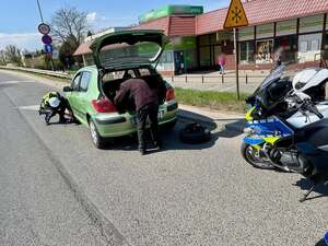 Mężczyzna stoi przy otwartym bagażniku pojazdu. Umundurowany policjant wymienia koło w samochodzie. W tle ruch uliczny i policyjne motocykle.