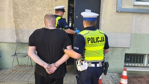 umundurowany funkcjonariusz Policji prowadzi zatrzymanego, a drugi funkcjonariusz z przodu otwiera drzwi