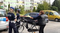 Dwie policjantki rozmawiają z rowerzystkę i wręczają jej kamizelkę odblaskową. W tle parking samochodowy i blok mieszkalny.