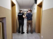 Policjant prowadzi zatrzymanego korytarzem Komendy Powiatowej Policji w Zgierzu na czynności. Mężczyźni są widoczni tyłem.