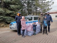 Na środku widoczny jest policyjny radiowóz przed którym leżą torby z prezentami i stoi dwóch policjantów w mundurach i jeden w ubraniu cywilnym (organizator przedsięwzięcia). Przed radiowozem stoi również trójka dzieci.