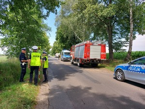 Miejsce wypadku. Radiowóz i wóz strażacki przy drodze. 3 policjantów w trakcie oględzin.