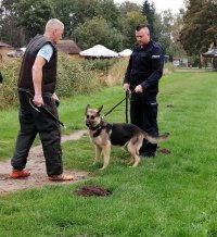 pozorant udający napastnika jest przeszukiwany przez policjanta i jednocześnie obserwowany przez psa, który stoi przed nim