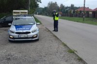 Policjant umundurowany z żółtą kamizelką z napisem policja stoi przy drodze obok radiowozu oznakowanego i mierzy prędkość.