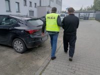 Policjant nieumundurowany, który ma założoną żółtą kamizelkę z napisem policja prowadzi obok idącego zatrzymanego mężczyznę do samochodu.