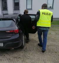 Policjant nieumundurowany, który ma założoną żółtą kamizelkę z napisem policja prowadzi obok idącego zatrzymanego mężczyznę do samochodu.