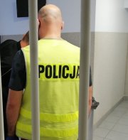 Nieumundurowany policjant z żółtą kamizelką z napisem policja stoi tyłem za okratowanymi drzwiami