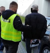 Nieumundurowany policjant w żółtej kamizelce prowadzi zatrzymanego mężczynę.