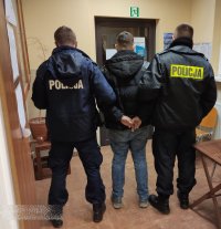 Umundurowani policjanci stoją tyłem z zatrzymanym mężczyzną, który stoi w środku.