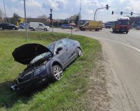 rozbity samochód koloru ciemnego stoi przewrócony w rowie na prawy bok, w pobliżu jest skrzyżowanie ulic i przejeżdżające samochody