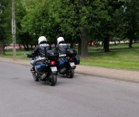 Dwaj umundurowani policjanci w białych kaskach na głowie jadą drogą siedząc na policyjnych motocyklach