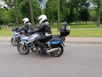 Dwaj umundurowani policjanci w białych kaskach na głowie jadą drogą siedząc na policyjnych motocyklach