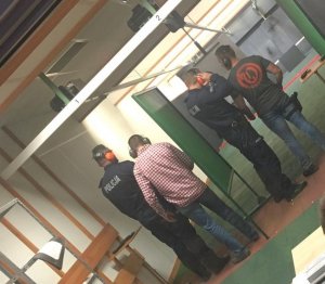 Policjanci w cywilnych ubraniach stoją obok umundurowanych policjantów, wszyscy mają nauszniki na uszach i są odwróceni tyłem. Stoją na stanowiskach przygotowanych do strzelania.