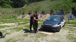 Dwóch policjantów w czarnych mundurach z nausznikami na uszach i ochronnych okularach z bronią w ręku stoi przed ostrzelanym radiowozem, obok stoją tarcze strzeleckie.