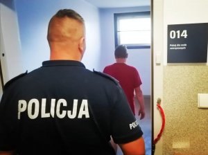 Policjant w granatowym mundurze odwrócony tyłem na plecach ma napis policja, stoi w drzwiach pomieszczenia gdzie stoi odwrócony tyłem zatrzymany mężczyzna w czerwonej koszulce.