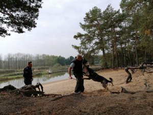 Umundurowany policjant stoi po lewej stronie zdjęcia i patrzy w kierunku psa atakującego mężczyznę w niebieskiej koszulce i pałką w ręku.