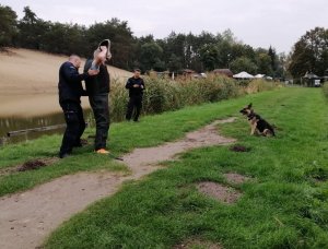 Umundurowani policjanci stoją obok mężczyzny trzymającego ręce w górze splecione na głowie, jeden z policjantów przeszukuje mężczyznę, na wprost którego siedzi pies i patrzy na niego.