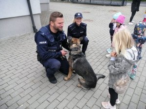 Umundurowany policjant kuca przy psie, obok niego umundurowana policjantka i wokół nich stoją dzieci.