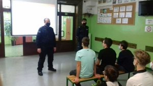 Policjant z policjantką stoją na środku sali i mówią do zwróconych w ich kierunku uczniów którzy siedzą przed nimi na ławkach.