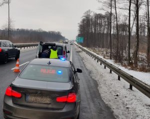 Nieoznakowany radiowóz ze świecącym na czerwono napisem policja który jest umieszczony za tylną szybą i z niebieskim światłem na dachu stoi na drodze przy miejscu zdarzenia drogowego, przed nim stoi policjant w żółtej kamizelce z innym mężczyzną przy zaparkowanym samochodzie przed radiowozem.