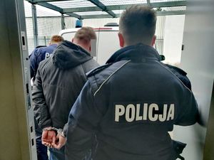 dwóch umundurowanych policjantów odwróconych tyłem prowadzi do radiowozu zatrzymanego mężczyznę, który stoi pośrodku policjantów