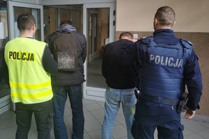 Od lewej stoi odwrócony tyłem policjant w żółtej kamizelce z napisem policja a przed nim stoi zatrzymany mężczyzna, obok nich stoi policjant w granatowym mundurze a przed nim stoi zatrzymany drugi mężczyzna.