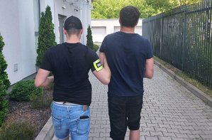 policjant z opaską na ramieniu prowadzi zatrzymanego mężczyznę