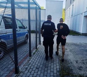 policjant prowadzi zatrzymanego mężczyznę, który ma założone kajdanki na rękach trzymanych z tyłu