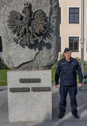 umundurowany policjant stoi przed budynkiem Wyższej Szkoły Policji w Szczytnie, po jego prawej stronie stoi symbol orła