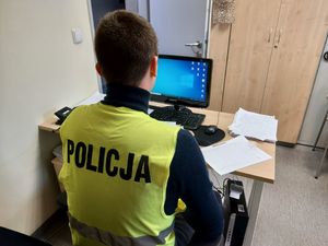policjant w żółtej kamizelce z napisem policja na plecach siedzi przy biurku na którym jest monitor komputera