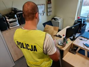 Policjant w żółtej kamizelce z napisem policja stoi odwrócony tyłem, w ręku trzyma kopertę