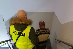 Dwóch policjantów, jeden z nich w żółtej kamizelce z napisem policja prowadzi zatrzymanego mężczyznę