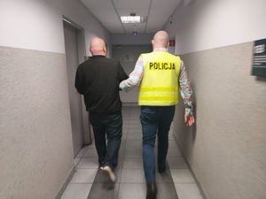 Policjant w żółtej kamizelce z napisem policja na plecach prowadzi zatrzymanego mężczyznę.