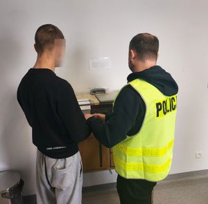 Zatrzymany mężczyzna stoi przy policjancie w żółtej kamizelce z napisem policja.