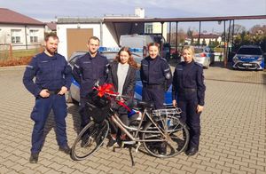 Dwóch policjantów w mundurach i dwie policjantki stoją obok młodej dziewczyny, która stoi w środku, przed nimi ustawiony rower z czerwoną kokardą, z tyłu stoją zaparkowane radiowozy.