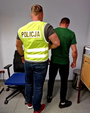 Policjant w żółtej kamizelce z napisem Policja prowadzi zatrzymanego mężczyznę.