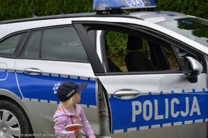 Policyjny radiowóz a przy nim mała dziewczynka odwrócona bokiem, na głowie ma założoną granatową czapkę z napisem policja.