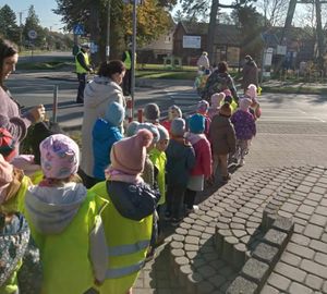 Policjanci stoją na drodze przez którą przechodzą dzieci w odblaskowych kamizelkach.