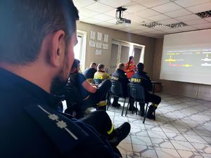 Policjant siedzi na sali wraz ze strażakami i patrzy w stronę rzutnika.