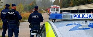 Trzech policjantów stoi obok radiowozu, w oddali są strażacy.