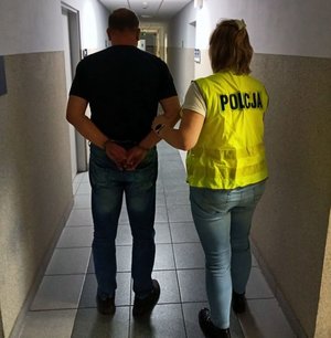 Policjantka w żółtej kamizelce z napisem policja prowadzi zatrzymanego mężczyznę.