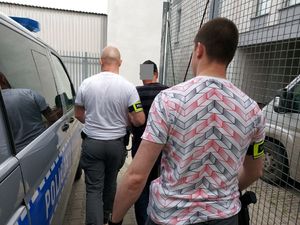 Dwóch policjantów z opaskami na przedramieniu z napisem policja prowadzi zatrzymanego mężczyznę.