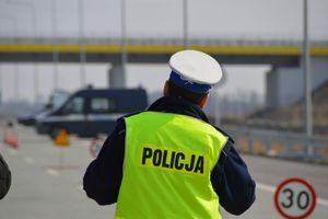 Policjant w białej czapce i żółtej kamizelce z napisem policja stoi na drodze.