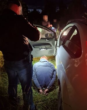 Policjant stoi przy samochodzie, przed nim na ziemi siedzi mężczyzna z kajdankami założonymi z tyłu na ręce.