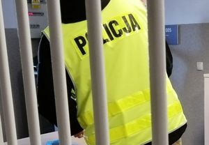 Policjant w kamizelce z napisem policja stoi za okratowanymi drzwiami.